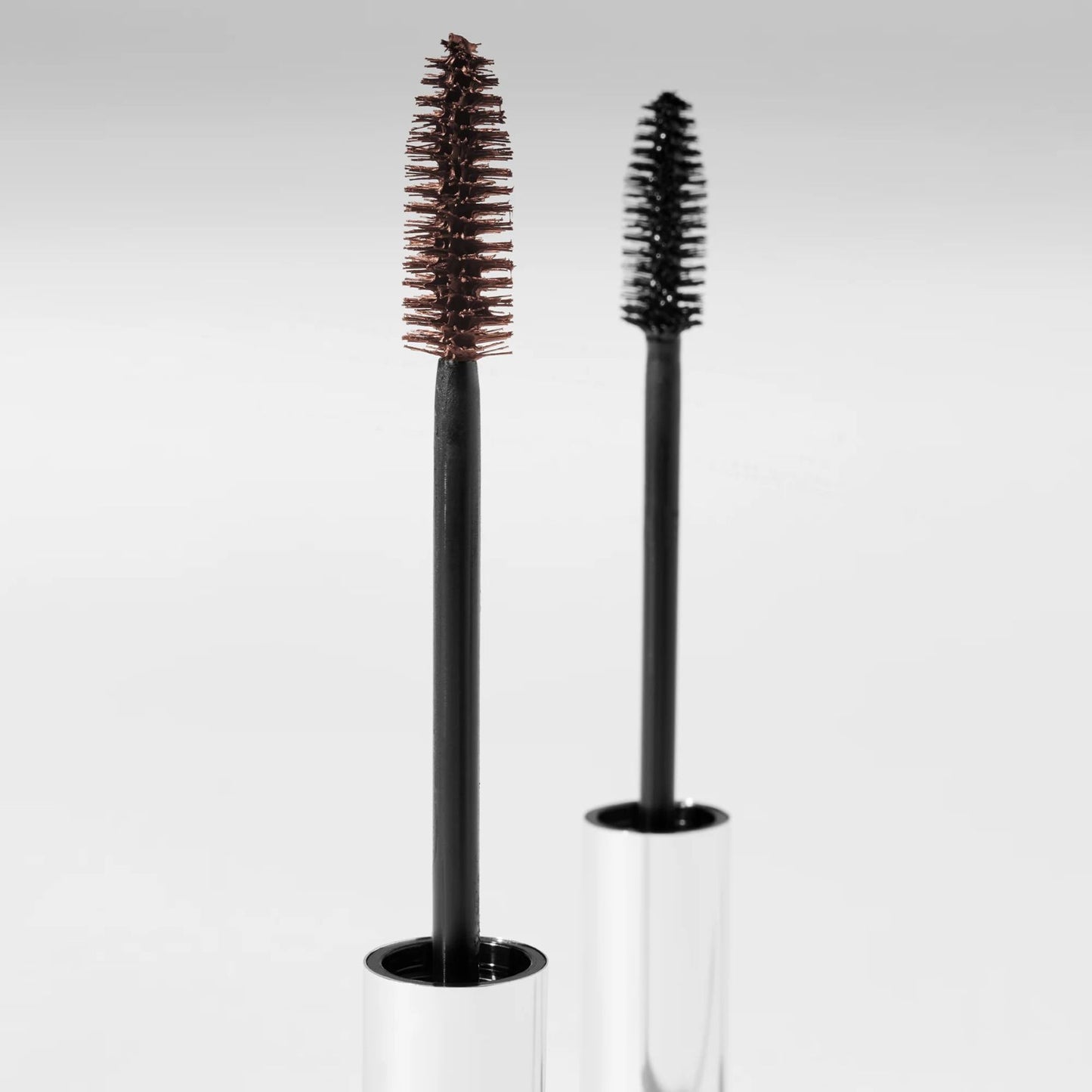 A black and brown serum mascara brushes with clear cylindrical holders on a light gray background